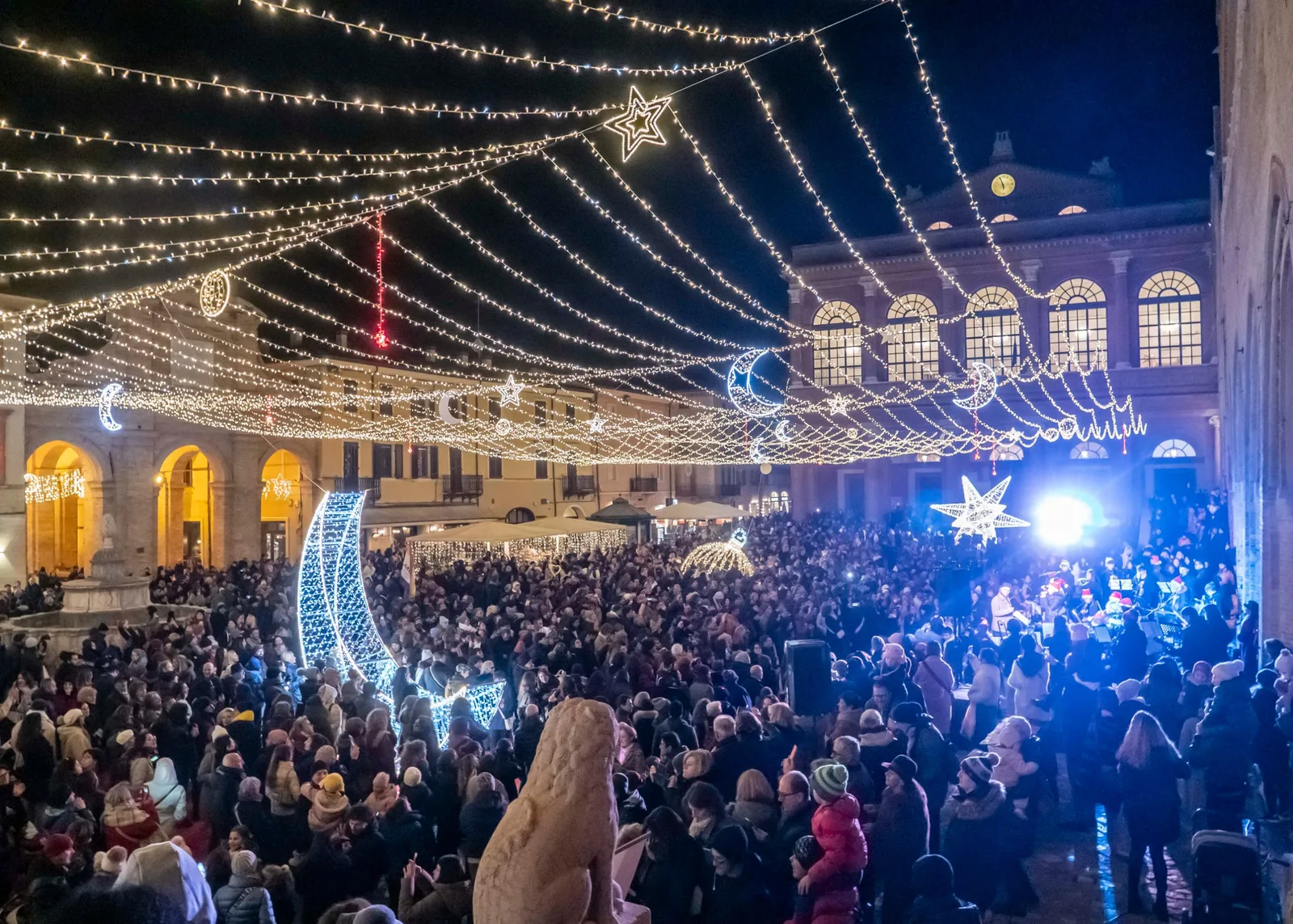 A Piazza Cavour emberekkel tele a karácsonyi díszkivilágítás alatt, világító holdakkal és csillagokkal, a Fontana della Pignával és a Teatro Gallival