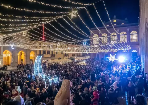 Piazza Cavour piena di gente sotto le luminarie di Natale, con le lune e le stelle luminose, la fontana della Pigna e il Teatro Galli
